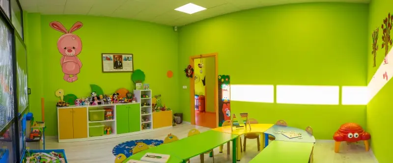 Yeşil duvarlı sınıfta alçak masalar, oyuncak rafları ve halı alanı